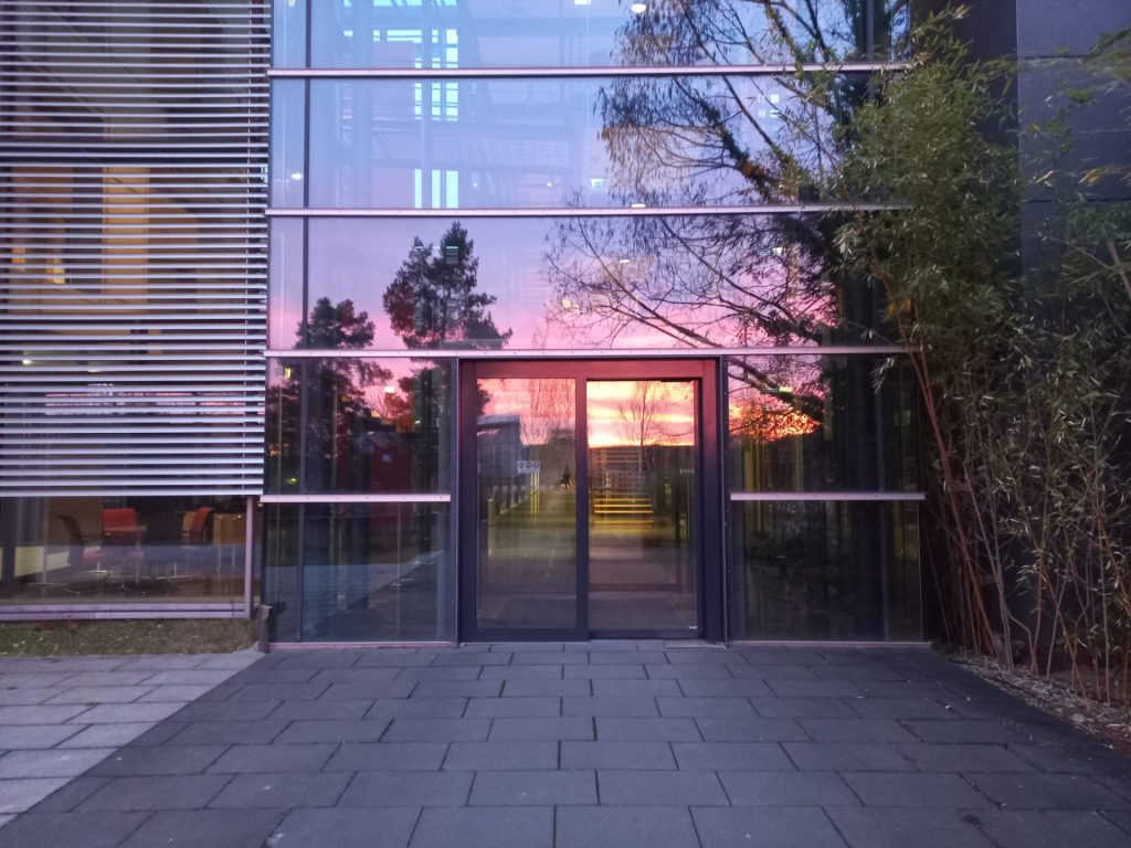 Foto der Eingangstür des Bioinstrumente-Zentrums. In der Glasfassade spiegelt sich ein bunter Sonnenaufgang.